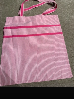 Rosa randig shoppingväska i bomull - Säljer en rosa shoppingväska med vita smala ränder och två breda axelband. Väskan har två dekorativa rosa band på framsidan och är tillverkad i mjuk bomull. Perfekt för att bära böcker, laptop eller snacks till plugget eller stan.