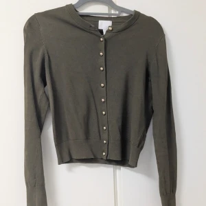 Olivgrön kofta från H&M med pärlknappar - Olivgrön kofta från H&M i mjuk bomullsblandning. Klassisk rund halsringning och långa ärmar. Snygga pärlknappar framtill ger en extra touch. Perfekt att slänga över en topp eller klänning för en chill look.