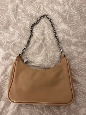 Beige axelväska med kedja - Snygg beige axelväska med silvrig kedjerem och dragkedja upptill. Väskan har en stilren, mjuk form och är tillverkad i syntetmaterial. Perfekt storlek för det viktigaste och har ett innerfack med dragkedja för extra förvaring.