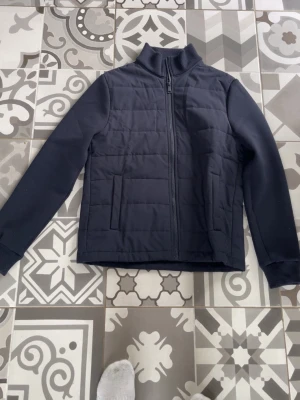 Svart quiltad vindjacka med hög krage - Stilren svart vindjacka med quiltad front och rygg, hög krage och hel dragkedja. Jackan har två sidofickor och släta ärmar med muddar. Materialet ser ut att vara syntet, perfekt för att hålla vinden borta. Passar dig som vill ha en enkel och modern look. Storlek TEEN S