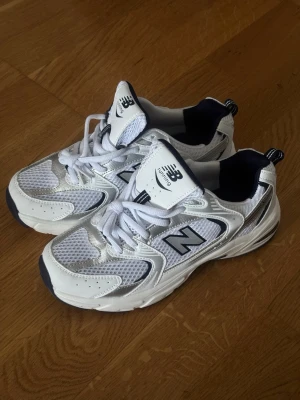 New Balance vita sneakers - Snygga vita New Balance sneakers med mesh och läderdetaljer. Skorna har klassisk N-logga på sidan, svarta och silverfärgade inslag samt en chunky sula med ABZORB-teknologi. Perfekta för dig som gillar retrostil och komfort.