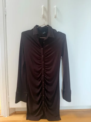 Brun klänning  - Mörkbrun skjortklänning från Gina Tricot med långa ärmar och knappar hela vägen framtill. Klänningen har en  rynkning längs knappraden och klassisk skjortkrage. Mjukt, stretchigt material som ger en bekväm passform.