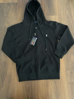 Svart hoodie från Polo Ralph Lauren - Snygg svart hoodie från Polo Ralph Lauren med dragkedja framtill, två fickor och klassisk huva med dragsnören. Liten vit logga broderad på bröstet. Tillverkad i mjukt bomullsmaterial som känns skönt mot huden. Perfekt för en avslappnad och stilren look.
