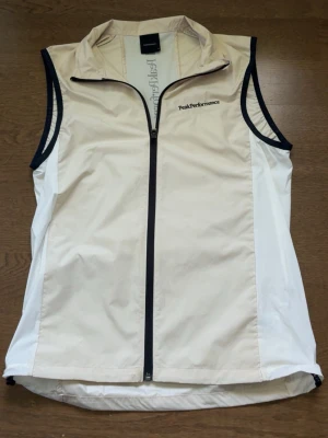 Beige väst från Peak Performance - Stilren, beige tunn väst från Peak Performance med svarta detaljer och dragkedja framtill. Västen har hög krage och logga på både bröstet och ryggen. Perfekt för lager-på-lager och passar till många olika outfits. Västen är riktigt fint skick och är perfekt till golf samt sommarväder.