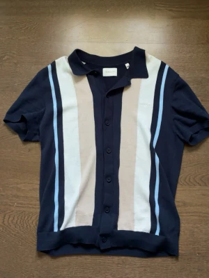 Mörkblå kortärmad skjorta från Gant - Snygg mörkblå kortärmad skjorta från Gant med knappar framtill. Nypris ligger på cirka 1600kr och tröjan är från förra sommarkollektionen av gant. Skjortan har vertikala ränder i beige, vitt och ljusblått på framsidan samt klassisk krage. Perfekt för en stilren och avslappnad look.