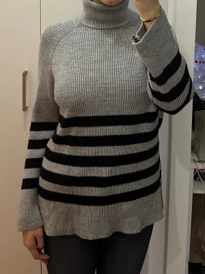 Grå stickad polotröja med svarta ränder - Säljer en grå stickad polotröja med breda svarta ränder på ärmar och kropp. Tröjan har hög polokrage, lång ärm och avslappnad passform. Ribbstickad struktur och mjukt material som känns skönt mot huden. Perfekt för kalla dagar.