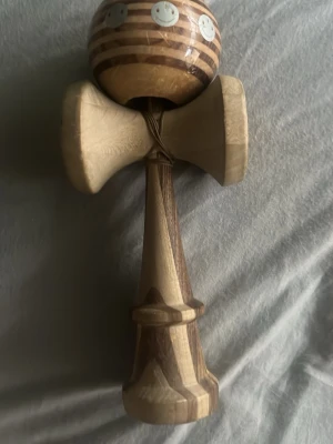 Kendama i trä med smiley-detaljer - Säljer en snygg kendama i trä med naturliga och mörkbruna ränder. Bollen har tre små smiley-ansikten i metall och hela leksaken har en cool, handgjord känsla. Perfekt för dig som gillar att trixa och utmana dig själv med klassiska spel.