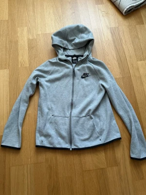 Grå zip hoodie från Nike - Snygg grå hoodie från Nike med dragkedja framtill och svart Nike-logga på bröstet. Hoodien har två fickor, huva och svarta detaljer vid ärmslut och nederkant. Tillverkad i mjukt bomullsmaterial som är skönt att bära.