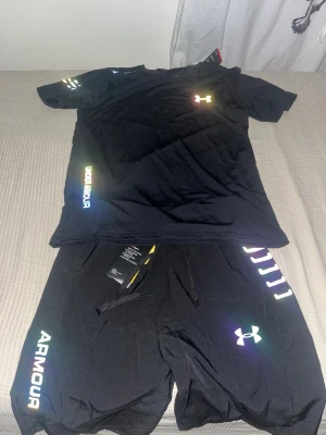 Svart träningsset från Under Armour - Snyggt svart träningsset från Under Armour med t-shirt och shorts. Setet har reflekterande loggor och textdetaljer i regnbågsfärger. Tillverkat i lätt och snabbtorkande material som passar perfekt för sommar och träning..