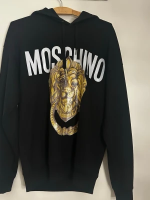 Svart Moschino hoodie med lejontryck - Svart hoodie från Moschino med stor vit logga och ett guldigt lejontryck på bröstet. Klassisk huva och ribbade muddar. Perfekt för dig som gillar statement-plagg och vill ha något unikt i garderoben.