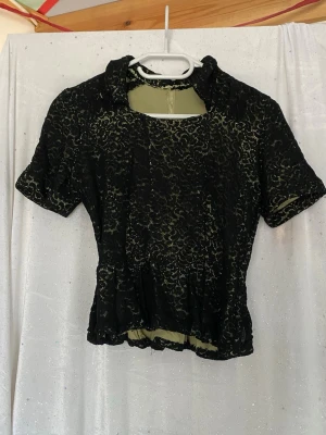 Svart spetsblus med kort ärm - Snygg svart spetsblus med korta ärmar och rund halsringning. Blusen har ett tunt grönt foder under spetsen och dragkedja på sidan som är lite sönder men funkar utan problem. Perfekt för dig som gillar en romantisk och vintage-inspirerad stil.