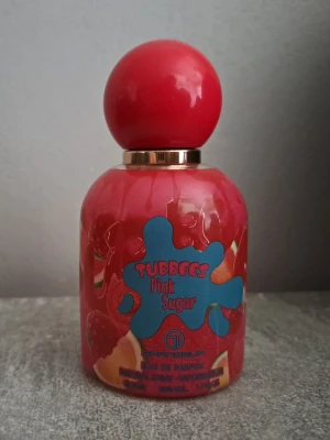 Tubbees Pink Sugar parfym - Tubbees Pink Sugar är en parfym i en rund, färgglad flaska med röd kork och guldigt halsband. Flaskan har en lekfull design med blå och röda detaljer samt fruktmotiv. Perfekt för dig som gillar söta och fruktiga dofter.
