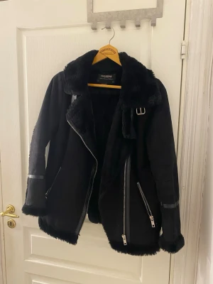 Svart pälsjacka från Pull&Bear - Snygg svart pälsjacka från Pull&Bear med mjukt foder, bred krage och dekorativa spännen. Jackan har dragkedjor framtill och vid fickorna samt detaljer i konstläder på ärmarna. Perfekt för kalla vinterdagar och ger en cool look. Pris kan diskuteras 
