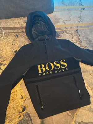 Svart hoodie från Hugo Boss - Snygg svart hoodie från Hugo Boss med stor gul logga på bröstet. Hoodien har dragkedjor på fickorna, mesh-detaljer på ärmarna och en stor huva. Perfekt för dig som vill ha en stilren och sportig look.