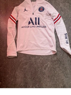 Vit långärmad PSG träningströja - Säljer en vit långärmad träningströja från PSG med Jordan-logga och klubbmärke på bröstet. Tröjan har dragkedja vid halsen, röda och svarta ränder på ärmarna samt tryck på ena ärmen och ryggen. Perfekt för fotbollsträning eller sport.
