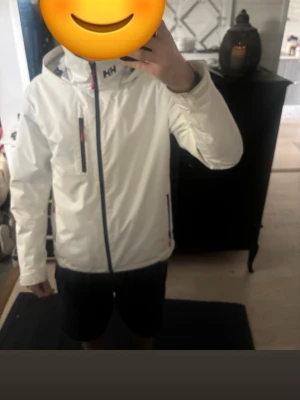 Vit vindjacka från Helly Hansen - Snygg vit vindjacka från Helly Hansen med svart dragkedja och röda detaljer på fickorna. Jackan har huva, hög krage och flera praktiska fickor. Perfekt för blåsiga dagar och enkel att matcha med olika outfits. Använd väldigt sällan skicket är så gott som nytt!