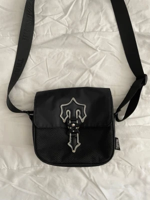 Svart axelväska från Trapstar - Snygg svart axelväska från Trapstar med justerbar rem och coolt emblem på locket. Väskan har en unik spänndetalj framtill och är gjord i slitstarkt tyg med meshdetaljer. Perfekt för att bära det viktigaste och ger en streetwear vibe. Pris kan diskutera
