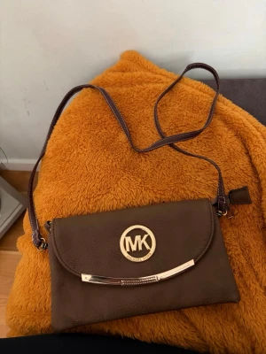 Brun axelväska från Michael Kors - Snygg brun axelväska från Michael Kors med guldfärgade detaljer och MK-logga framtill. Väskan har ett smidigt format, justerbar axelrem och dragkedjefack på baksidan. Tillverkad i konstläder som ger en stilren look.