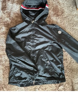 Svart vindjacka från Moncler - Snygg svart vindjacka från Moncler med huva och dragkedja framtill. Jackan har Moncler-logga på ärmen och vita samt röda detaljer på huvan. Tillverkad i ett glansigt, lätt material som står emot vind. Perfekt för dig som vill ha en stilren och sportig look.