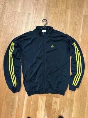 Adidas träningsjacka - Snygg mörkblå/mörkgrön träningsjacka från Adidas med gula ränder längs ärmarna och Adidas-logga på bröstet. Jackan har dragkedja framtill, ståkrage och två sidofickor. Tillverkad i ett lätt och sportigt material, perfekt för träning eller chill.