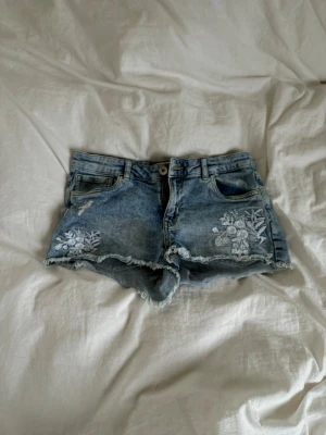 Blå lågmidjade denimshorts med broderi - Säljer ett par blå denimshorts med råa kanter och snyggt vitt blommigt broderi på framsidan. Shortsen har klassiska fickor fram och bak samt knapp och dragkedja. Dom är lågmidjade och passar bra. Dom går även att justera i midjan 