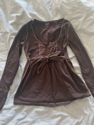 Hollister babydoll top med knytning  - Brun Hollister topp. Storlek S. Mycket bra skick. Säljer för jag inte använder den. Köpt på Vinted. Priset kan diskuteras Man kan ha knytningen både fram eller bak!