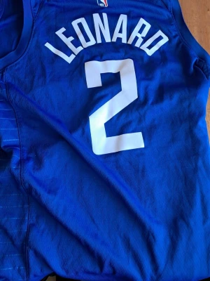 Nike NBA LA Clippers Kawhi Leonard #2 Swingman Jersey, Icon Edition.  - Säljer ett blått basketlinne från NBA med tryck 'LEONARD' och nummer 2 på ryggen samt 'CLIPPERS' på framsidan. Linnet är ärmlöst och har klassisk sportdesign, perfekt för basketträning eller att bära som supporter. Materialet är lätt och andas.