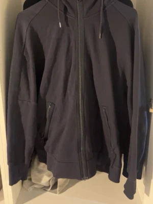 Mörkblå hoodie med dragkedja - Snygg mörkblå hoodie med dragkedja framtill och två fickor med dragkedja. Tröjan har huva med snörning och coola detaljer som glasögon i huvan. Perfekt för dig som gillar streetwear och vill ha något unikt i garderoben.