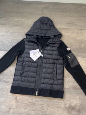Svart Moncler jacka med huva - Säljer en svart Moncler jacka med quiltad front och huva. Jackan har stickade ärmar med en ficka och Moncler-logga på ena armen. Dragkedja framtill och två sidofickor. Materialet är en kombination av nylon och stickat tyg. Passar även storlek S