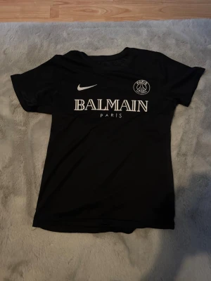 Svart PSG x Balmain t-shirt Nike - Svart t-shirt med Paris Saint-Germain och Balmain Paris-tryck på bröstet. Klassisk rund halsringning och korta ärmar. Snygg sportig stil med vit logga och text framtill. Perfekt för dig som gillar streetwear och fotboll.
