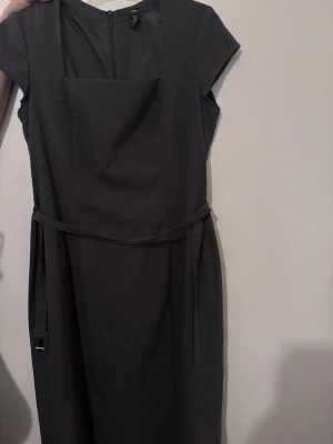 Mörkgrå klänning med bälte H&M - Stilren mörkgrå klänning från H&M med korta ärmar och fyrkantig urringning. Klänningen har ett avtagbart bälte i midjan och dragkedja bak. Perfekt för dig som gillar en klassisk och enkel look.