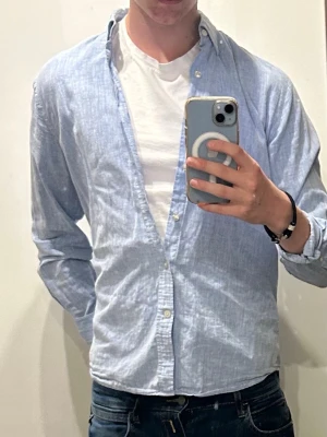 Ljusblå oxfordskjorta - Säljer en klassisk ljusblå oxfordskjorta med button-down krage och vita knappar. Skjortan har lång ärm och en avslappnad passform, perfekt att bära öppen över en t-shirt. Materialet är bomull och ger en fräsch och stilren look.