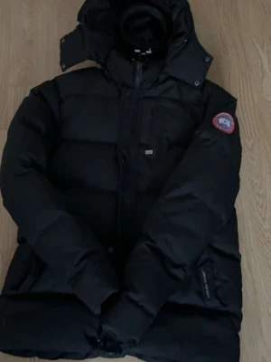 Svart dunjacka från Canada Goose - Säljer en svart dunjacka från Canada Goose med huva och klassisk patch på ärmen. Jackan har en puffig design, dragkedja och fickor framtill. Perfekt för kalla vinterdagar och riktigt varm tack vare dunfyllningen.