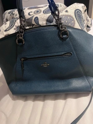 Blå handväska i läder från Coach - Snygg blå handväska från Coach i äkta läder med grova silverfärgade kedjedetaljer och dragkedja framtill. Väskan har både handtag och axelrem, samt en praktisk ytterficka med Coach-logga. Perfekt för dig som vill ha en stilren och rymlig väska.
