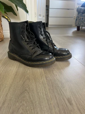 Svarta Dr. Martens kängor i läder - Klassiska svarta Dr. Martens kängor i äkta läder med snörning och den ikoniska gula sömmen runt sulan. Robust design med högt skaft och grov sula som ger en cool och tidlös look. Perfekta för dig som vill ha en edgy stil.