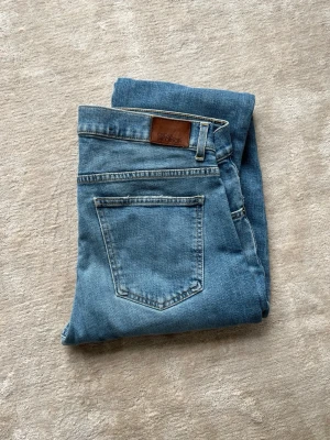 Blå jeans från Oscar Jacobson - Snygga blå jeans från Oscar Jacobson i klassisk rak modell. Jeansen har fem fickor, ljusblå tvätt och detaljer i brunt läder vid linningen. Perfekta för en avslappnad och stilren look.