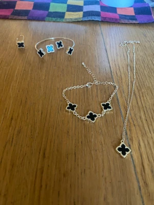 Smyckesset med svarta fyrklöver - Säljer ett snyggt smyckesset med armband, halsband, örhängen och ring i guldfärgad metall. Alla delar har svarta fyrklöverformade hängen med guldfärgad kant. Perfekt för dig som gillar stilrena och trendiga accessoarer.