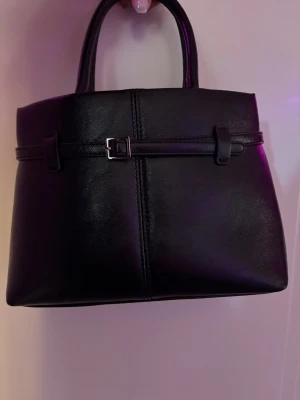 Svart handväska med bältdetalj - Stilren svart handväska i slätt konstläder med två handtag och snygg bältdetalj framtill. Väskan har en fyrkantig form och minimalistisk design som passar till många olika outfits. Perfekt för dig som gillar klassiska accessoarer med en modern twist. I väldigt fint skick och kommer med ett längre band. 