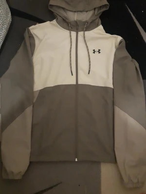 Under Armour beige vindjacka med huva - Snygg vindjacka från Under Armour i beige och ljusgrått med dragkedja framtill, justerbar huva och elastiska muddar. Jackan har meshfoder och logga på bröstet. Perfekt för blåsiga dagar och sportiga outfits.
