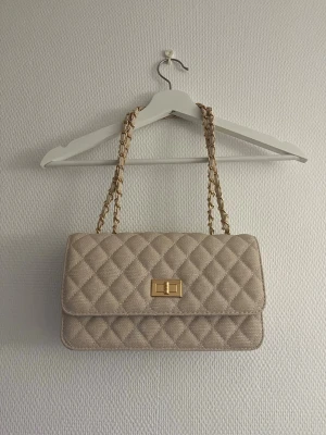 Beige quiltad handväska med kedja - Säljer en beige quiltad handväska med guldiga kedjor som axelband och ett snyggt vridlås i guld på framsidan. Väskan har en rektangulär form och är tillverkad i ett textilmaterial med diamantmönster. Perfekt för dig som gillar klassisk och elegant stil.