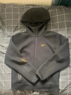 Svart Nike Tech Fleece hoodie - Snygg svart Nike Tech Fleece hoodie med dragkedja och huva. Har gul Nike-logga på bröstet och texten ACG på ärmen. Två fickor med dragkedja framtill. Perfekt för en sportig och avslappnad stil.