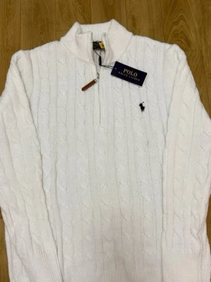 Vit kabelstickad tröja Polo Ralph Lauren - Kabelstickad vit tröja från Polo Ralph Lauren med halv dragkedja och hög krage. Tröjan har det klassiska broderade logotypen på bröstet och ribbstickade muddar vid ärmslut och nederkant. Perfekt för en stilren och tidlös look.
