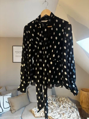 Svart prickig blus med scarf - Trendig svart blus från NA-KD med vita prickar och långa, puffiga ärmar. Blusen har ett elegant knytband/scarf vid halsen och är tillverkad i ett lätt, transparent material som ger en luftig känsla. Perfekt för dig som gillar en cool och chic stil. Strl 32 men passar större!