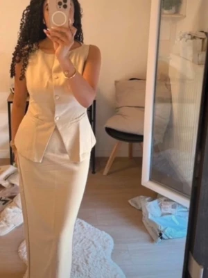 Topp och kjol set  - Stilren beige kostym med ärmlös kavaj och matchande lång kjol. Kavajen har markerad midja, knappar framtill och klassisk krage. Perfekt för dig som gillar en elegant och clean look.Sjukt snyggt elegant sett som är helt ny och oanvänd. Storlek M. Kostym material.