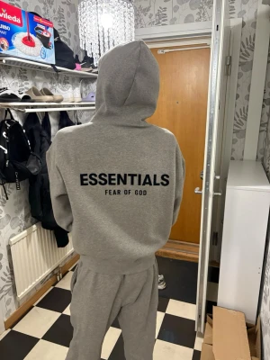 Grå Essentials Fear of God hoodie - Säljer en grå hoodie från Fear of God Essentials med stor svart logga på ryggen. Hoodien har en klassisk passform, huva och ribbade muddar. Perfekt för dig som gillar streetwear och vill ha en stilren look.