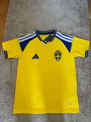 Sverige fotbollströja Adidas gul/blå - Gul fotbollströja från Adidas med blå detaljer och Sveriges landslagsemblem på bröstet. Tröjan har korta ärmar, blåa ränder på axlarna och V-ringad krage. Tillverkad i lätt och ventilerande material med Climalite-teknologi.