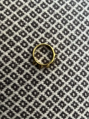 Guldfärgad ring med nitar - Cool ring i guldfärgad metall med flera spetsiga nitar runt hela ringen. Perfekt för dig som gillar edgy och unik stil. Ringen har en rund form och sticker verkligen ut med sina detaljerade nitar.