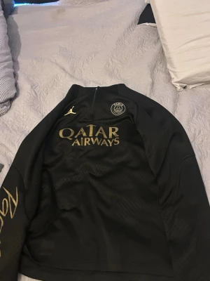 Svart PSG träningströja Jordan - Säljer en svart träningströja från PSG x Jordan med guldfärgade detaljer och Qatar Airways-tryck på bröstet. Tröjan har halv dragkedja, PSG-logga och Jordan-logga framtill. Perfekt för fotbollsträning eller chill.