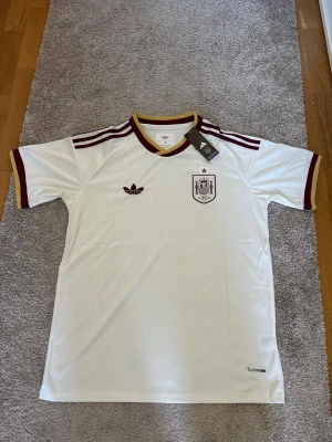 Vit Spanien Adidas fotbollströja - Säljer en vit Spanien fotbollströja från Adidas med vinröda och gula detaljer. Tröjan har korta ärmar, tre klassiska Adidas-ränder på axlarna och Spaniens landslagsemblem på bröstet. Tillverkad i lätt och ventilerande Climalite-material.