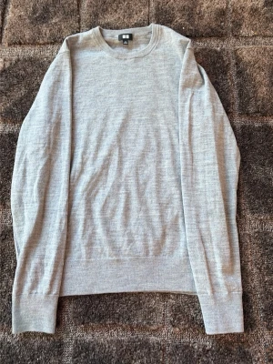 Ljusgrå stickad tröja från Uniqlo - Ljusgrå stickad tröja från Uniqlo med rund halsringning och långa ärmar. Tröjan är tillverkad i mjukt material och har ribbade muddar vid hals, ärmslut och nederkant. Perfekt för lager-på-lager och enkel att matcha med jeans eller chinos.